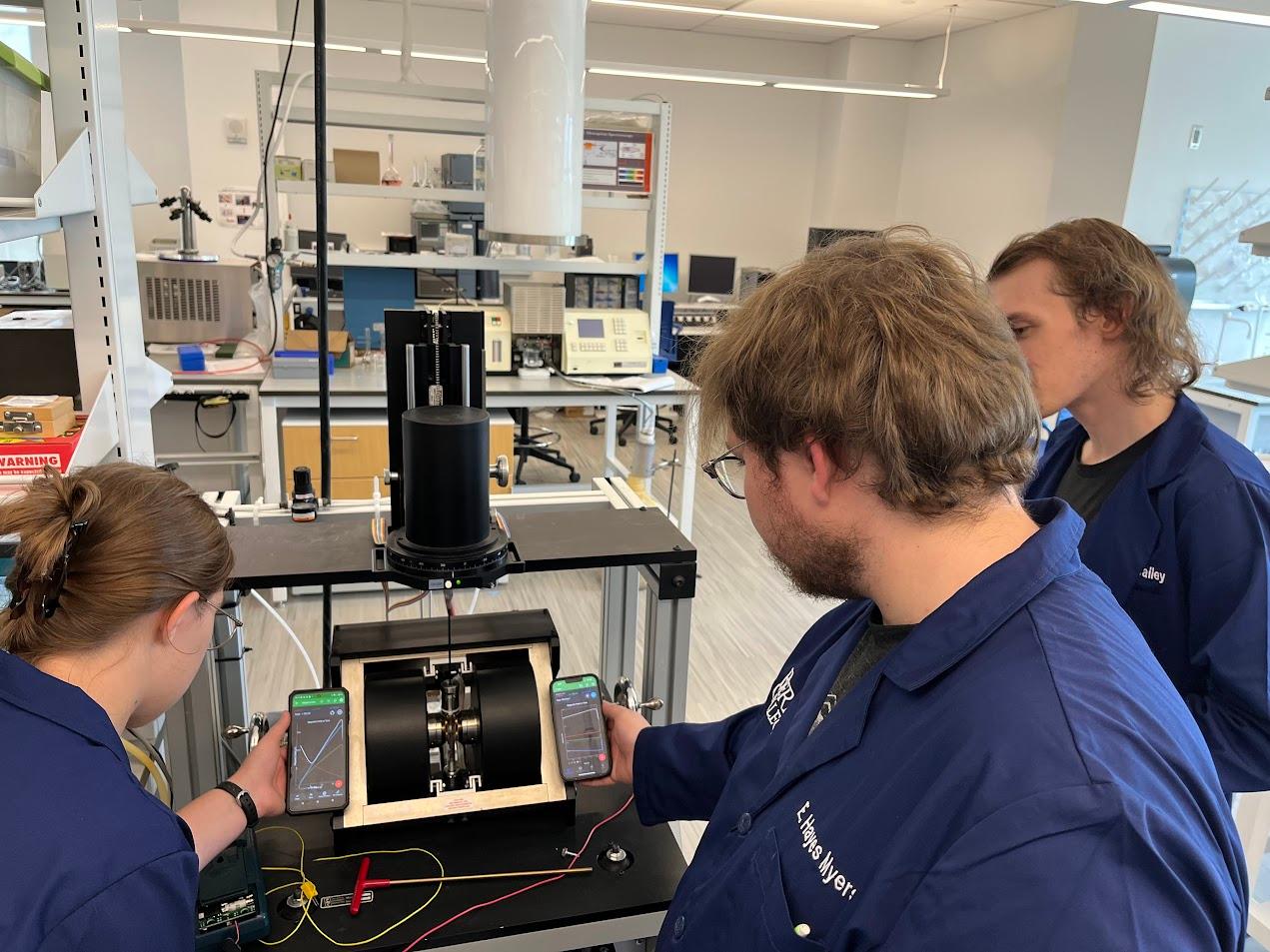 Three Berea College physics students wearing personalized lab coats look and gesture towards a machine in a lab.