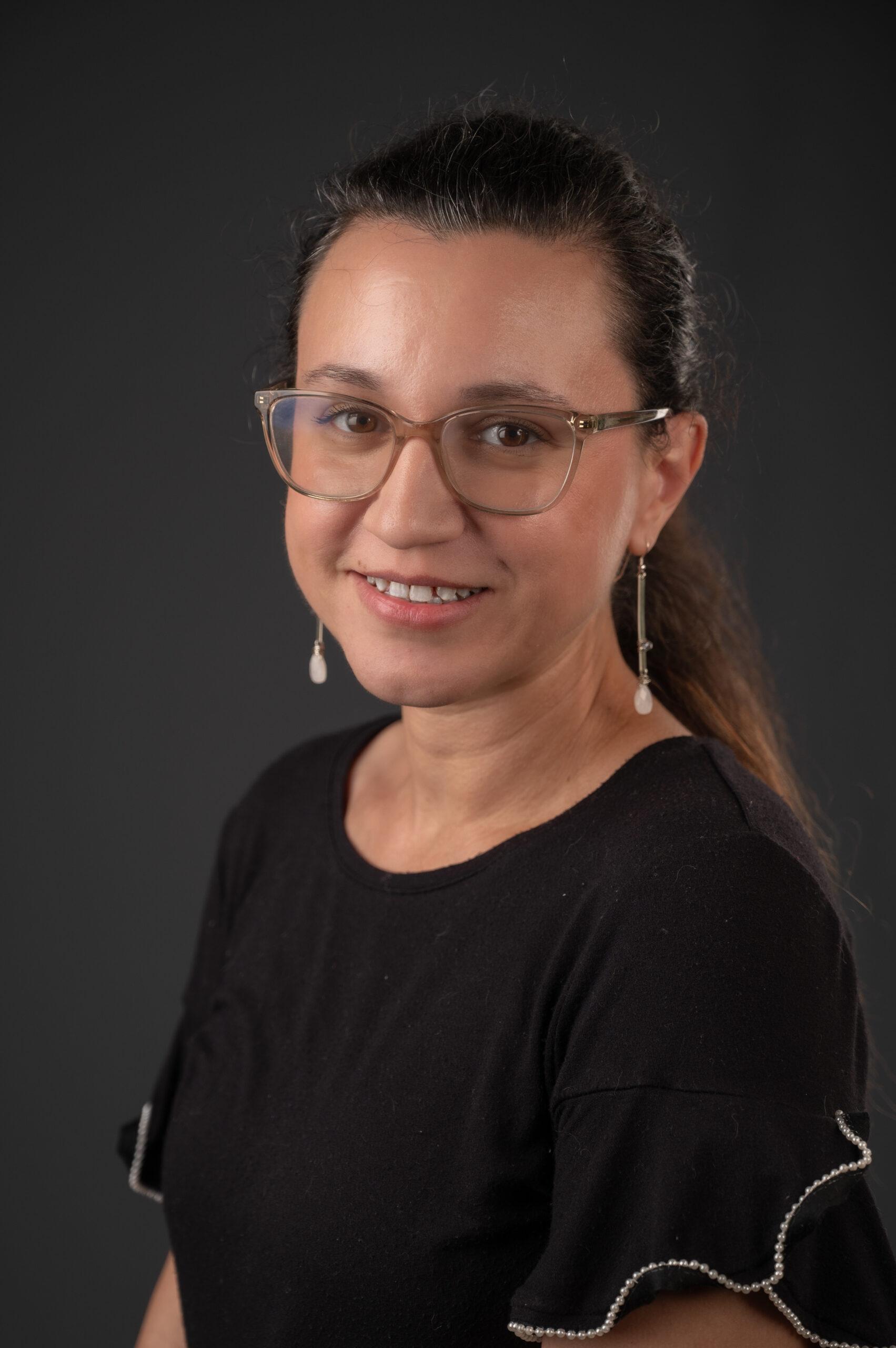 Jenny Boyle Akins headshot, sitting. Behind is a grey background and is wearing black smiling