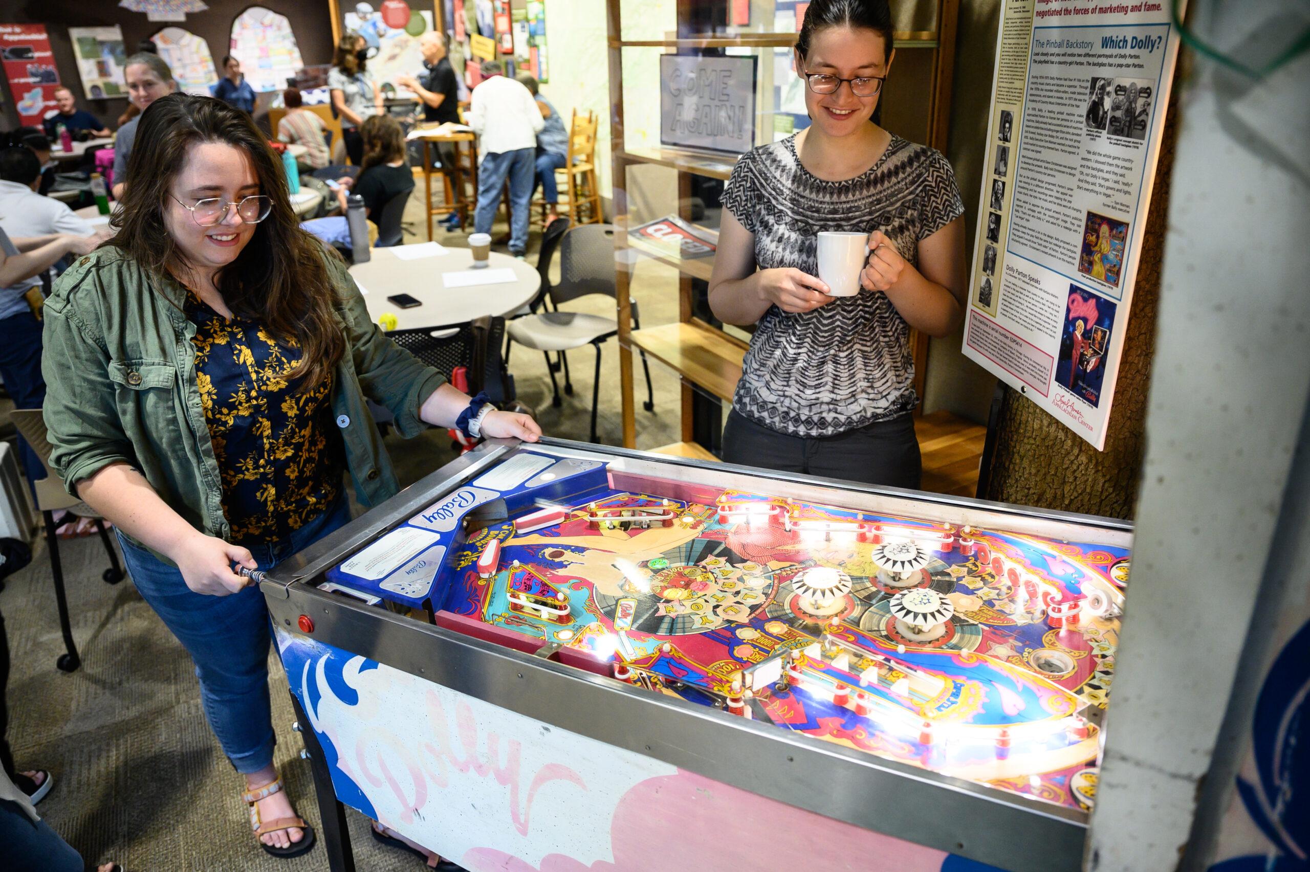 One woman enjoys a hot drink in the Loyal Jones Appalachian Center, while watching another person plays pinball on the infamous Dolly Parton pinball machine located in the Center.