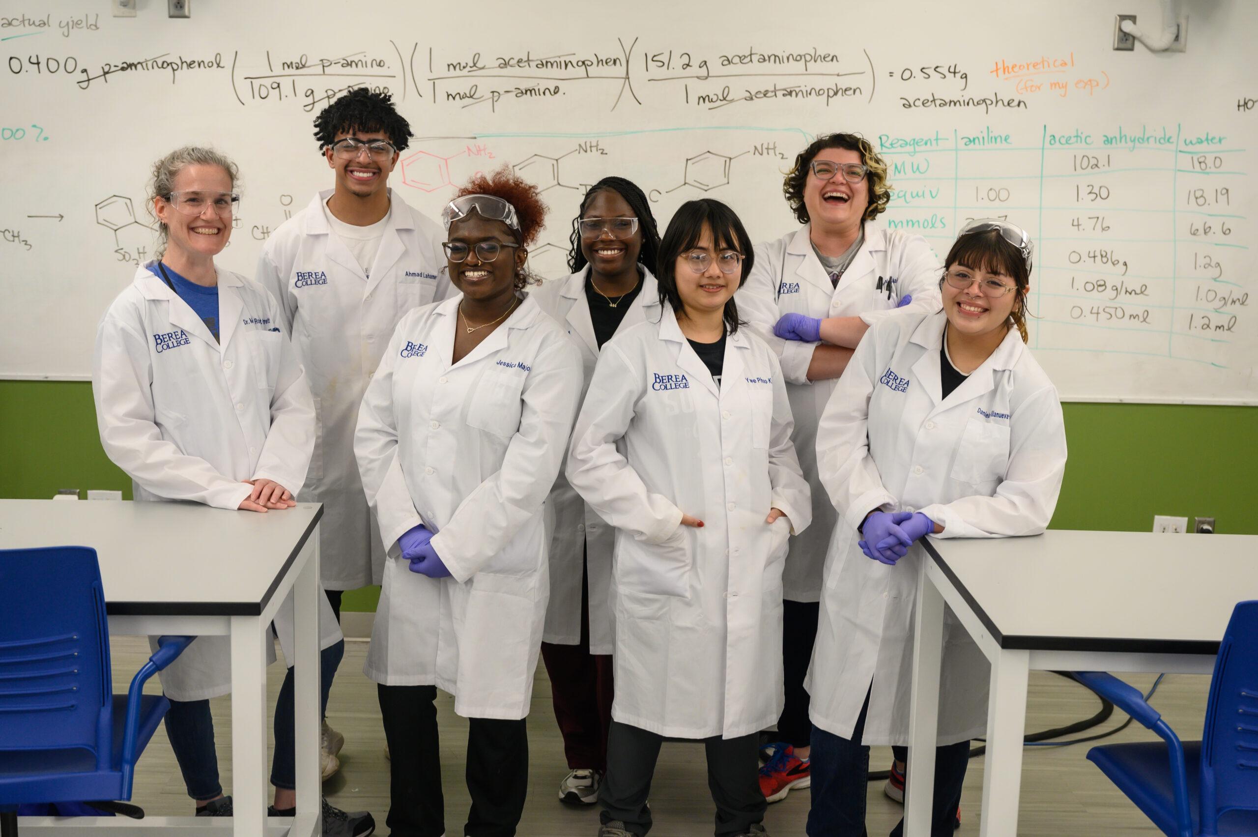 A professor and a group of students standing in lab coats and purple gloves smiling at the camera. There is a whiteboard in the background with text written on it.
